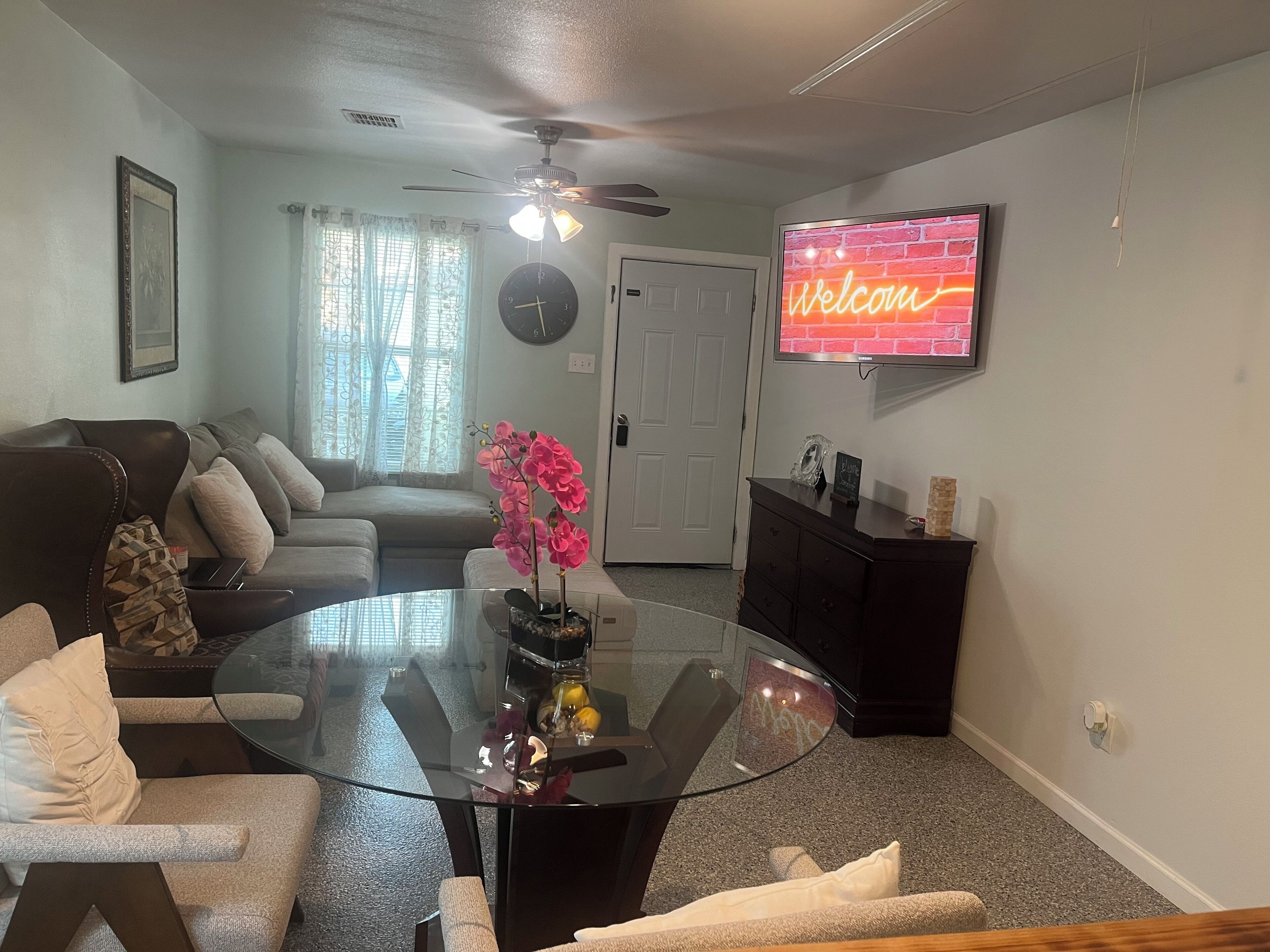 Una sala de estar con una mesa de comedor de vidrio, un sofá, un televisor y un reloj en la pared.