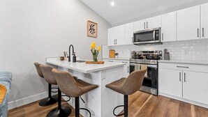 Een moderne keuken met witte kasten, een centraal eiland met barstoelen, een magnetron, fornuis en een spoelbak.