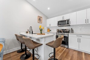 A modern kitchen with white cabinetry, a central island with bar stools, a microwave, stove, and a sink. - 2BR Garden Suite, U of A, Whyte Ave, Rogers Place, AC - Garage Parking (EDMONTON)