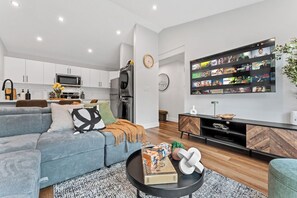 A modern living room with a grey sofa, a coffee table with books, a TV wall-mounted above a black cabinet, and a kitchen area in the background. - 2BR Garden Suite, U of A, Whyte Ave, Rogers Place, AC - Garage Parking (EDMONTON)