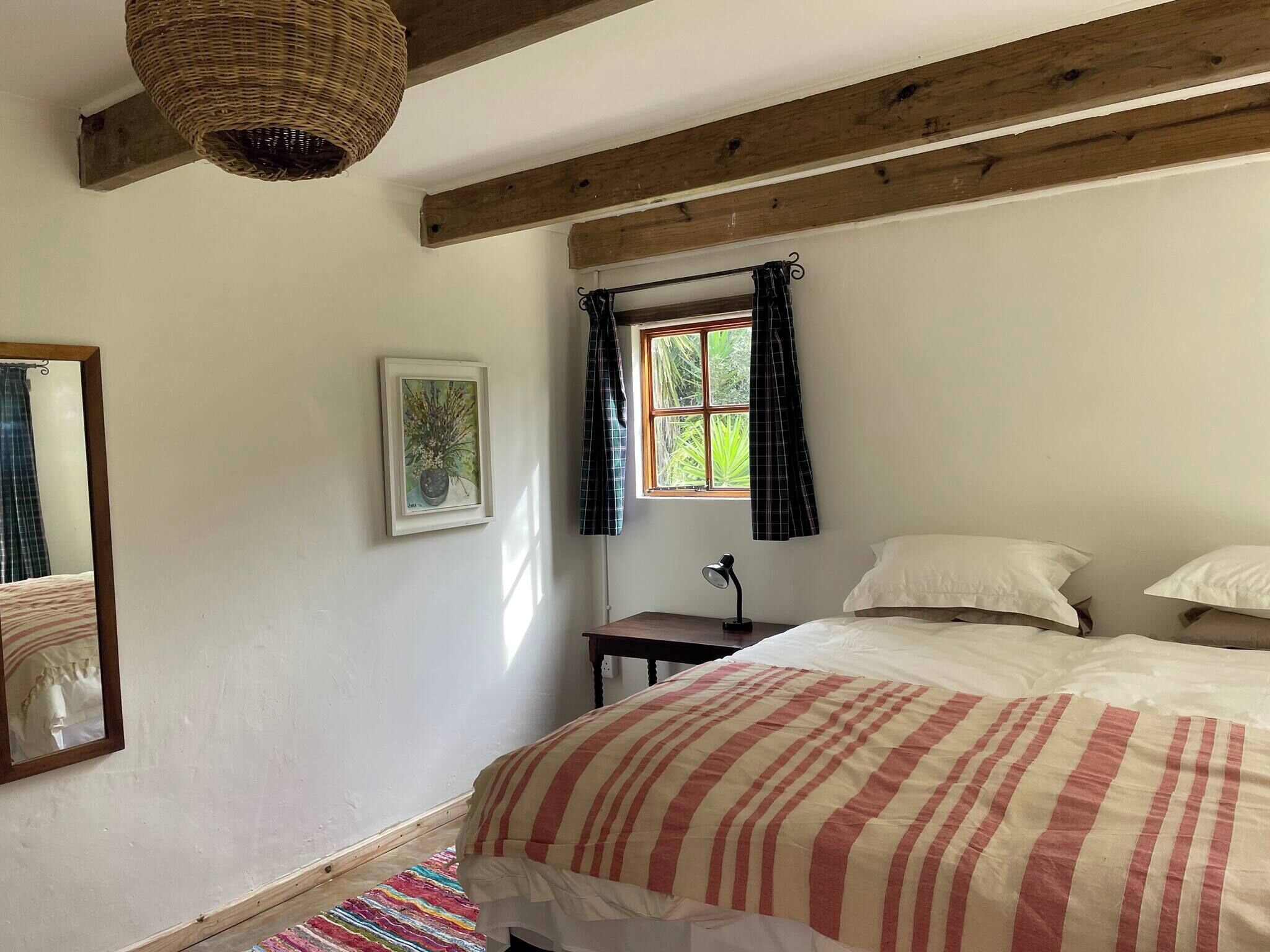 Un dormitorio con cama, ventana con cortinas, mesita de noche, lámpara, un cuadro enmarcado en la pared y un espejo.