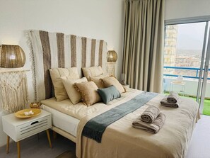 Ein Hotelzimmer mit einem großen Bett, einem Nachttisch, einer Lampe und Blick auf ein Gebäude durch das Fenster.