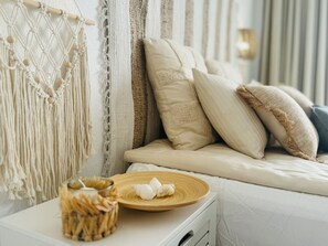 Ein Bett mit weißer Bettwäsche und mehreren Kissen, ein Wandbehang aus Makramee und ein kleiner Tisch mit einem Korb und einer Platte mit einer Kerze.