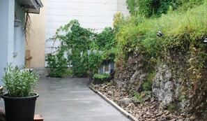 A narrow alleyway with a concrete path, a white wall, and dense greenery including ivy and various plants. - Vacation apartment Hexenhäusla Peace and relaxation in the heart of Franconian Switzerland (Gößweinstein)