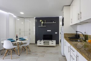 A modern kitchen with white cabinets, a granite countertop, a built-in TV, and a dining area with a table and chairs. - Luxury Summerland: Stylish 1BR Retreat in Los Cristianos with Pool & Terrace (Los Cristianos)