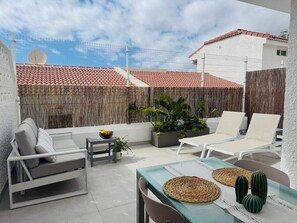 Eine Terrasse mit einem weißen Sofa, zwei Liegestühlen, einem Tisch mit gewobenen Unterlagen und Topfpflanzen.