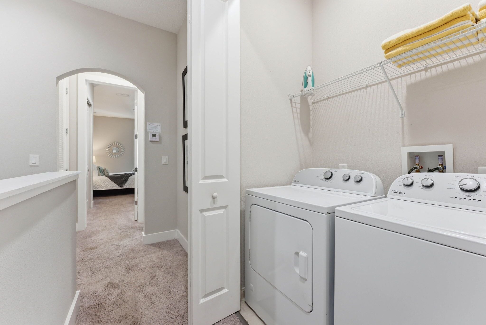 A laundry room with two white front-loading washers and dryers, a drying rack with yellow towels, and a hallway leading to a bedroom.
