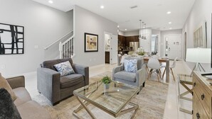 A modern living room with a glass coffee table, grey sofas, and a staircase leading to a kitchen area. - Family Retreat With 4bed Suites & Private Jacuzzi (Kissimmee)