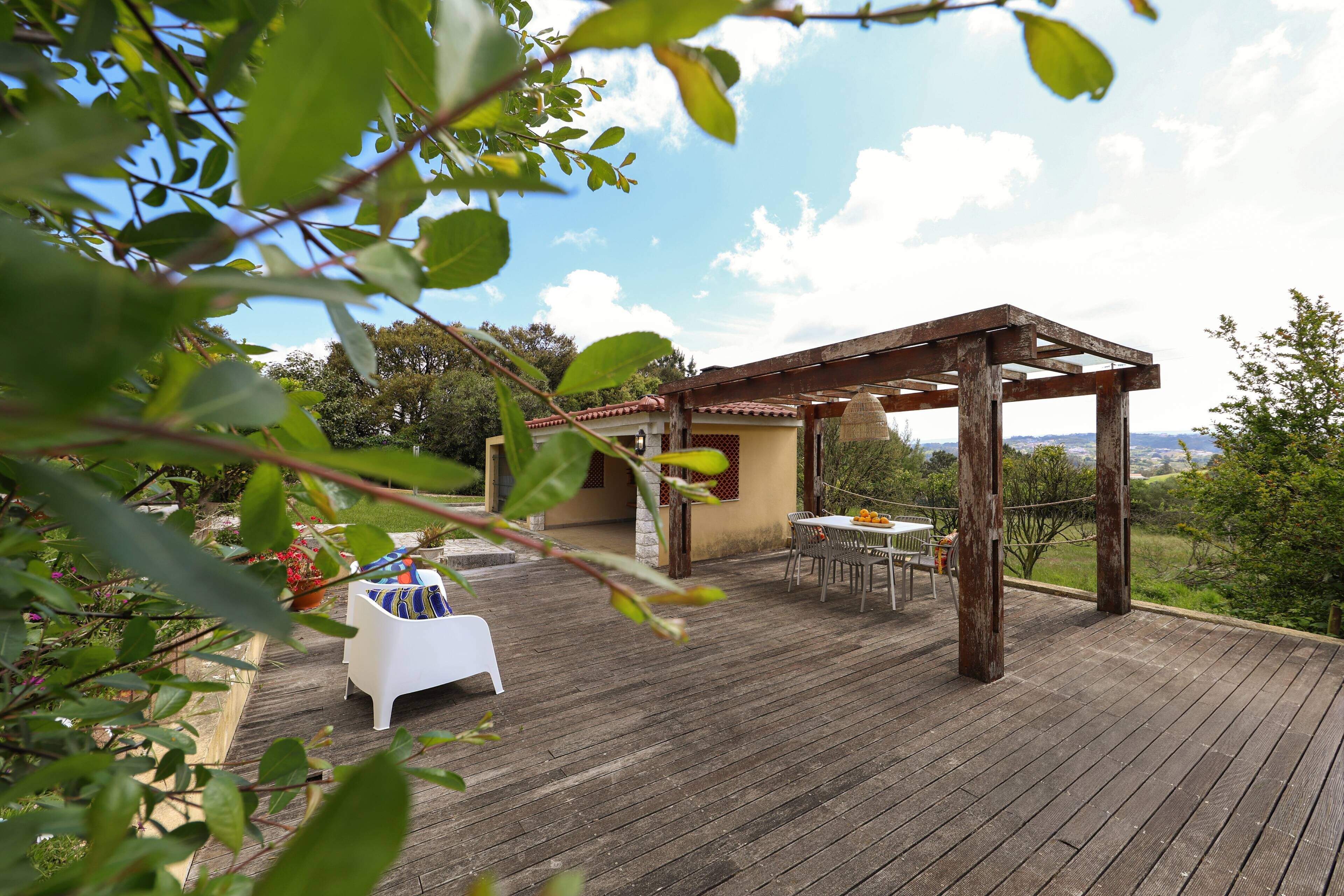 A wooden deck with a pergola, a white table with chairs, and a view of the countryside.