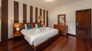 A hotel room with a large bed, a bedside table with a lamp, a dresser, a mirror, and a door. - Casa Favorita - Rustic Hideaway (Alcobaça)