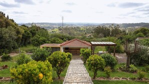 A house with a stone pathway leading to a garage, surrounded by lush greenery and a view of distant hills. - Casa Favorita - Rustic Hideaway (Alcobaça)