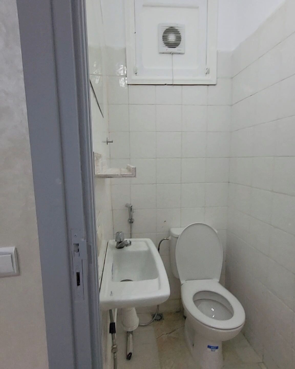 Ein kleines, sauberes Badezimmer mit Toilette, Waschbecken und Wandlüfter.