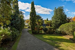 A gravel pathway leading through a lush garden with various trees and shrubs, a clear sky, and a distant view of a house. - Okawa Bay Waterfront | Lakefront with Jetty (Rotorua)