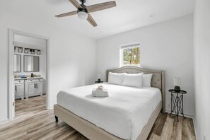 Ein Schlafzimmer mit Bett, Deckenventilator, Fenster, Nachttisch und Blick auf eine Küche.