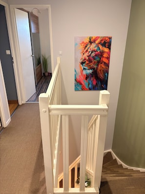 Un escalier avec une rampe blanche, un tableau représentant un lion et un couloir menant à une autre pièce.