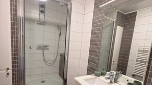 Et badeværelse med en brusekabine med glasvægge, en hvid vask med spejl og mørke fliserede vægge.