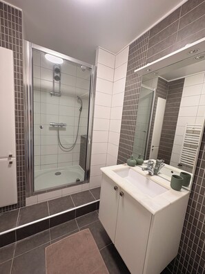 Une salle de bain avec une douche à parois de verre, un lavabo blanc avec un miroir et des murs recouverts de carreaux foncés.