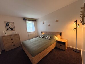 Une chambre à coucher avec un lit en bois, des tables de chevet, un radiateur, une fenêtre avec des rideaux, des œuvres d’art murales et une lampe sur pied.