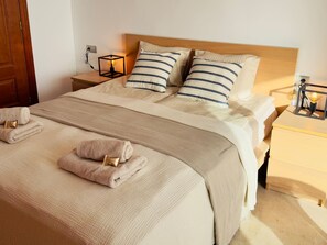 A neatly made bed with striped pillows, a beige blanket, and two folded towels with wooden clips. There are bedside tables with lamps and a wooden headboard. - Casa Heidi - Pool, Garage & Terrace - Puerto de Santiago (Santiago del Teide)