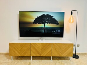 A flat-screen TV on a white stand with a wooden cabinet below, a black floor lamp to the right, and a router on the stand. - Casa Heidi - Pool, Garage & Terrace - Puerto de Santiago (Santiago del Teide)