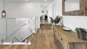 Un interior moderno con una escalera, piso de madera, barandilla de cristal, una consola de madera y un elemento decorativo en la pared.