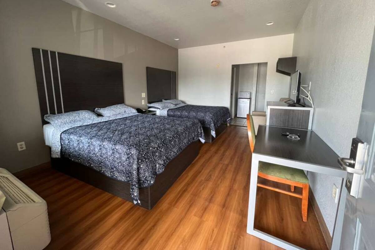A modern hotel room with two beds, a desk, and a kitchenette.