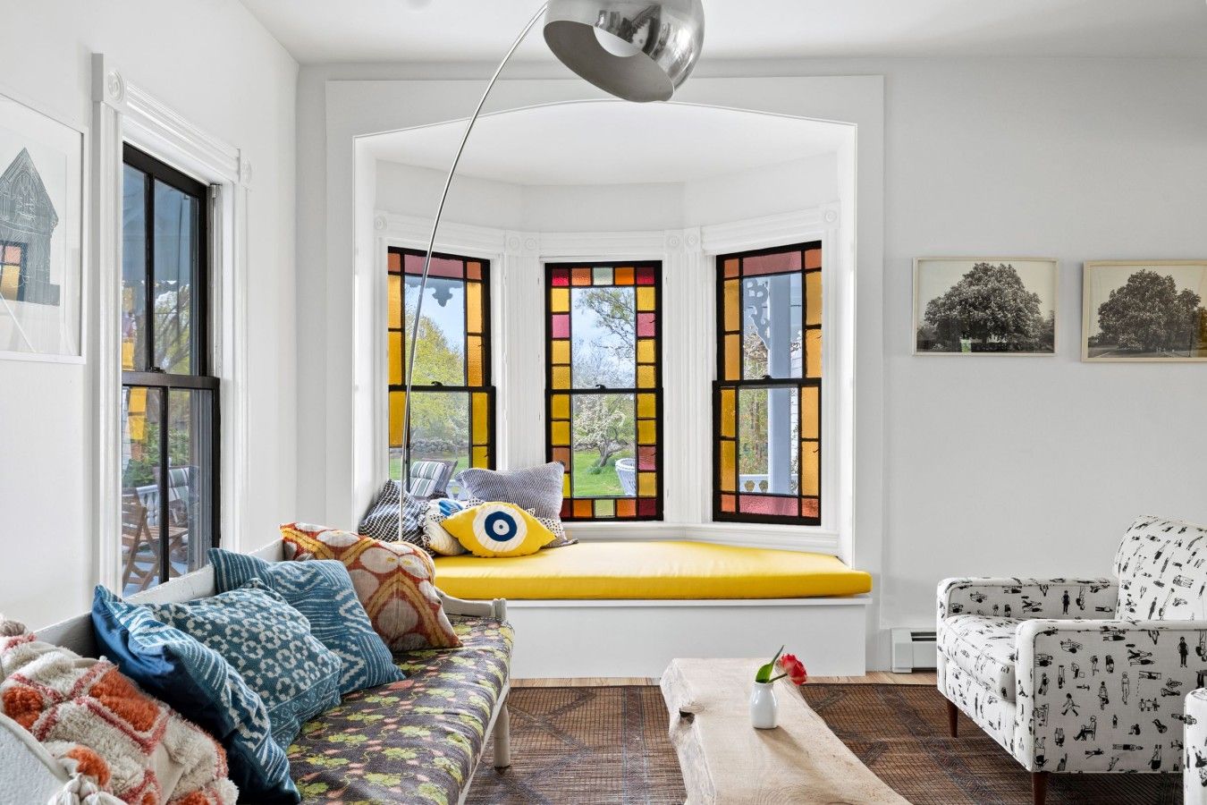 A cozy living room with a bay window, a sofa with patterned cushions, a patterned armchair, and a vase with a flower.