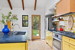 A modern kitchen with a black countertop, yellow cabinets, a stainless steel refrigerator, and a wooden door leading to a garden.