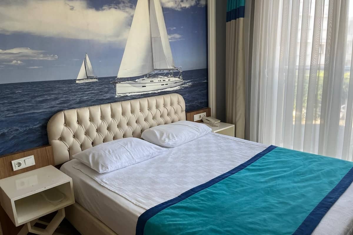 A hotel room with a bed, a nightstand, a wall mural of sailboats, and a window with curtains.