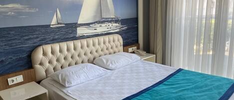 A hotel room with a bed, a nightstand, a wall mural of sailboats, and a window with curtains.