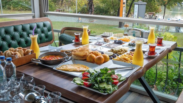 A breakfast buffet with various dishes and drinks, a green upholstered bench, and a view of a lake and trees. - River Suite Otel (Manavgat)