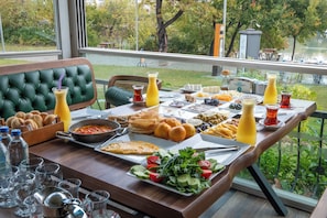 Un bufé de desayuno con diversos platos y bebidas, un banco tapizado de verde y vistas a un lago y árboles.