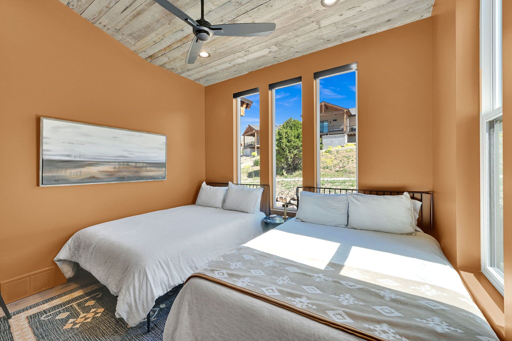A bedroom with two beds, a ceiling fan, large windows, and a painting on the wall.