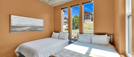 A bedroom with two beds, a ceiling fan, large windows, and a painting on the wall.