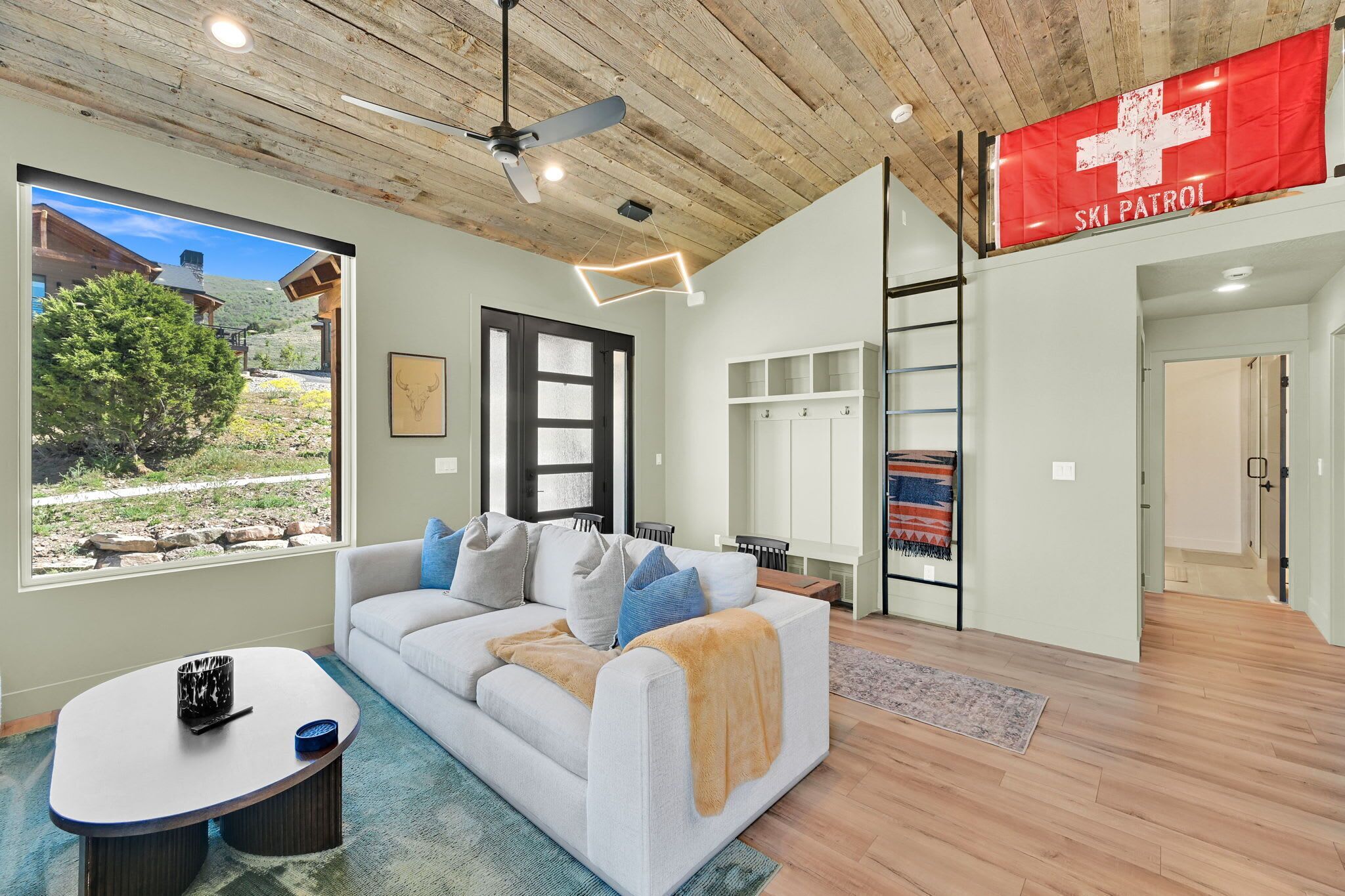 A modern living room with a white sofa, a coffee table, and a ceiling fan. There is a large window with a view of a mountain and a ski patrol sign.