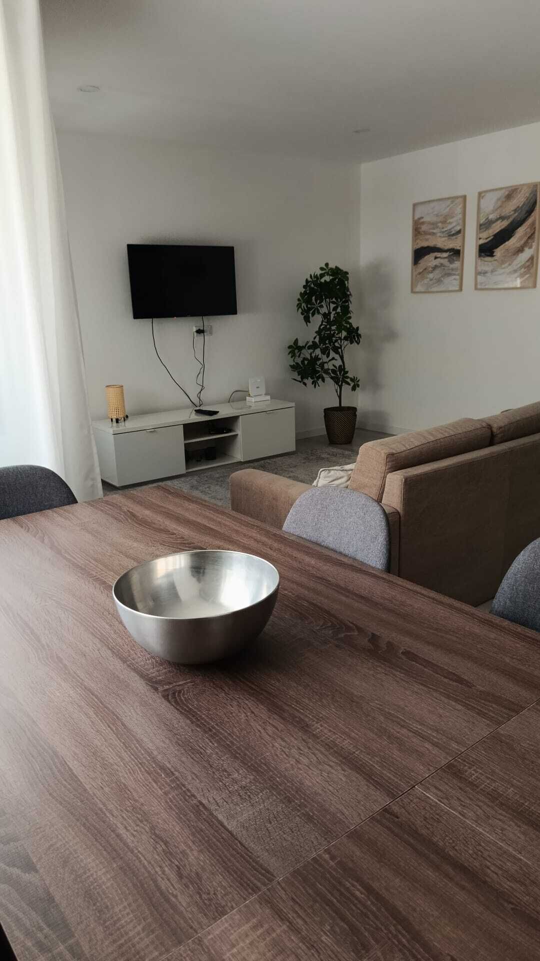 A modern living room with a wooden dining table, a sofa, a TV mounted on the wall, and a potted plant.