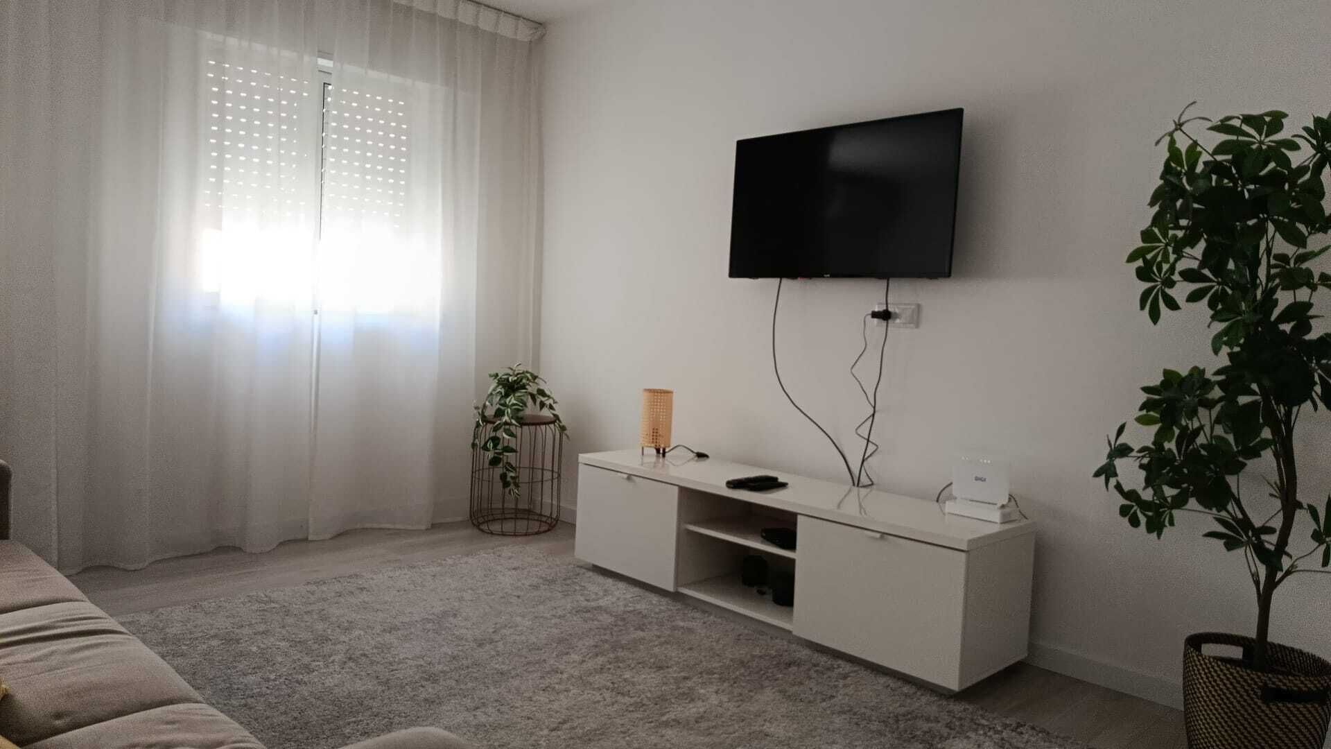 A living room with a flat-screen TV mounted on the wall, a white entertainment unit, a sofa, and a potted plant.
