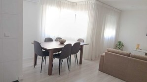 Et spiseområde med et træbord og stole, en skål, og en sofa i et lyst rum med hvide vægge og et stort vindue.