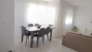 Un comedor con una mesa y sillas de madera, un cuenco y un sofá en una habitación luminosa con paredes blancas y un ventanal grande.