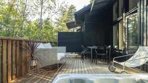 A wooden deck with a hot tub, outdoor seating, and a large window overlooking a forest.