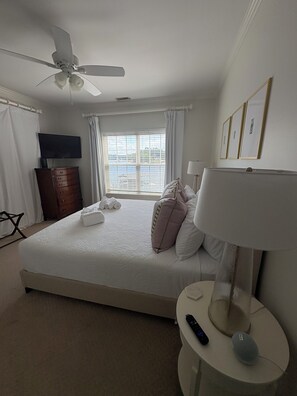 Une chambre à coucher avec un lit, un ventilateur de plafond, une table de chevet avec une lampe, une télécommande et une fenêtre avec des rideaux.