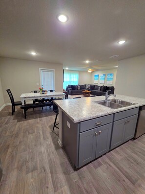 A modern kitchen with a central island, a dining area with a table and chairs, and a living room with a sofa and a ceiling fan. - Spacious 4 bedroom sleeps 10 one level house only minutes to the beach! (Foley)