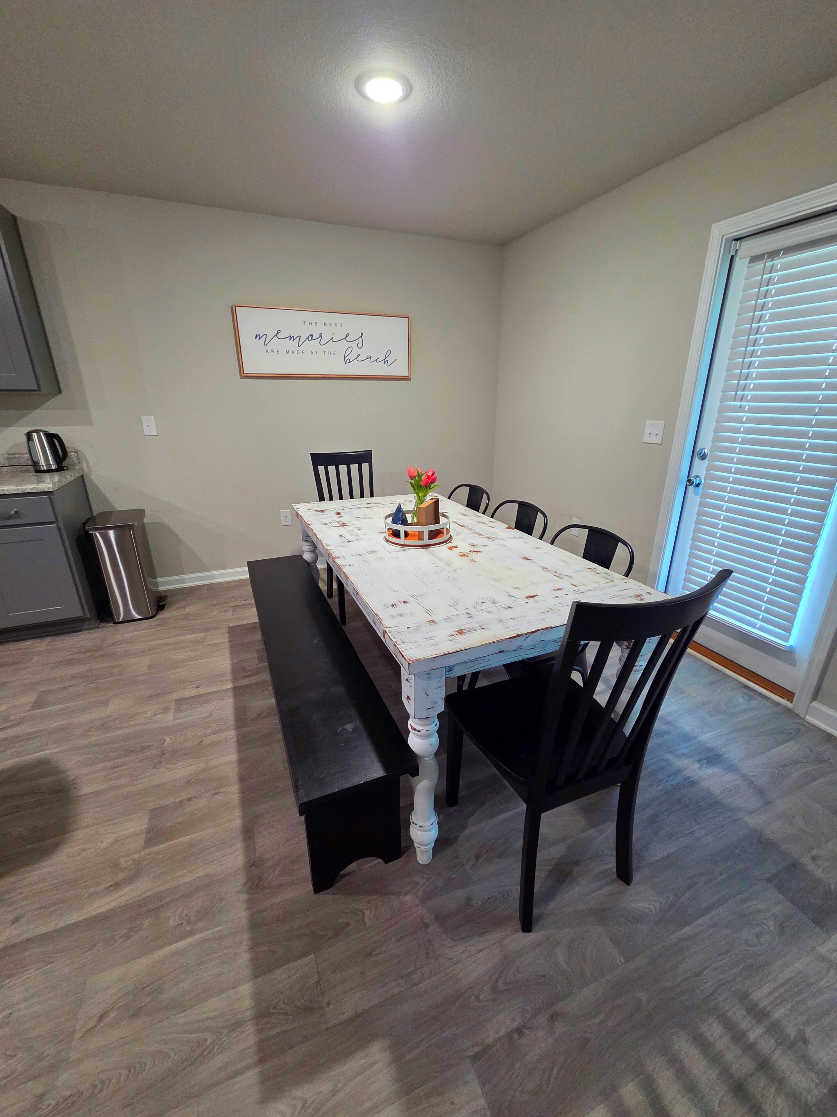 A dining area with a table, chairs, a bench, a kitchen area with a kettle, and a wall with a framed quote.