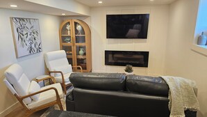 Ein Wohnzimmer mit schwarzem Ledersofa, einem weißen Sessel, einem Holzschrank, einem Flachbildfernseher und einem Kamin.