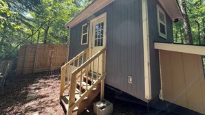 Une petite construction en bois d’un seul étage, dotée d’une porte d’entrée et d’une petite fenêtre, entourée d’une clôture en bois et d’arbres.