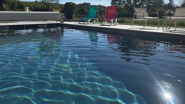 Et klart pool med synlige vandringe, omgivet af liggestole og et træ i baggrunden.