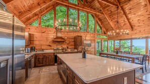 A spacious kitchen with a large island, wooden ceiling beams, and large windows overlooking a forest. - Luxury Lodge*Stunning Views*Prime-Location*Arcade (Sevierville)