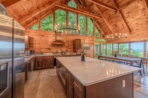 A spacious kitchen with a large island, wooden ceiling beams, and large windows overlooking a forest. - Luxury Lodge*Stunning Views*Prime-Location*Arcade (Sevierville)