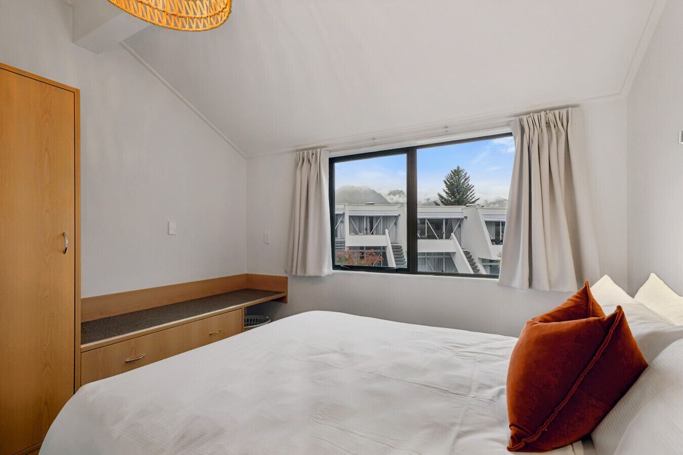 Ein Schlafzimmer mit einem Bett, einem Holzschrank, einer Bank und einem Fenster mit Blick auf Gebäude.