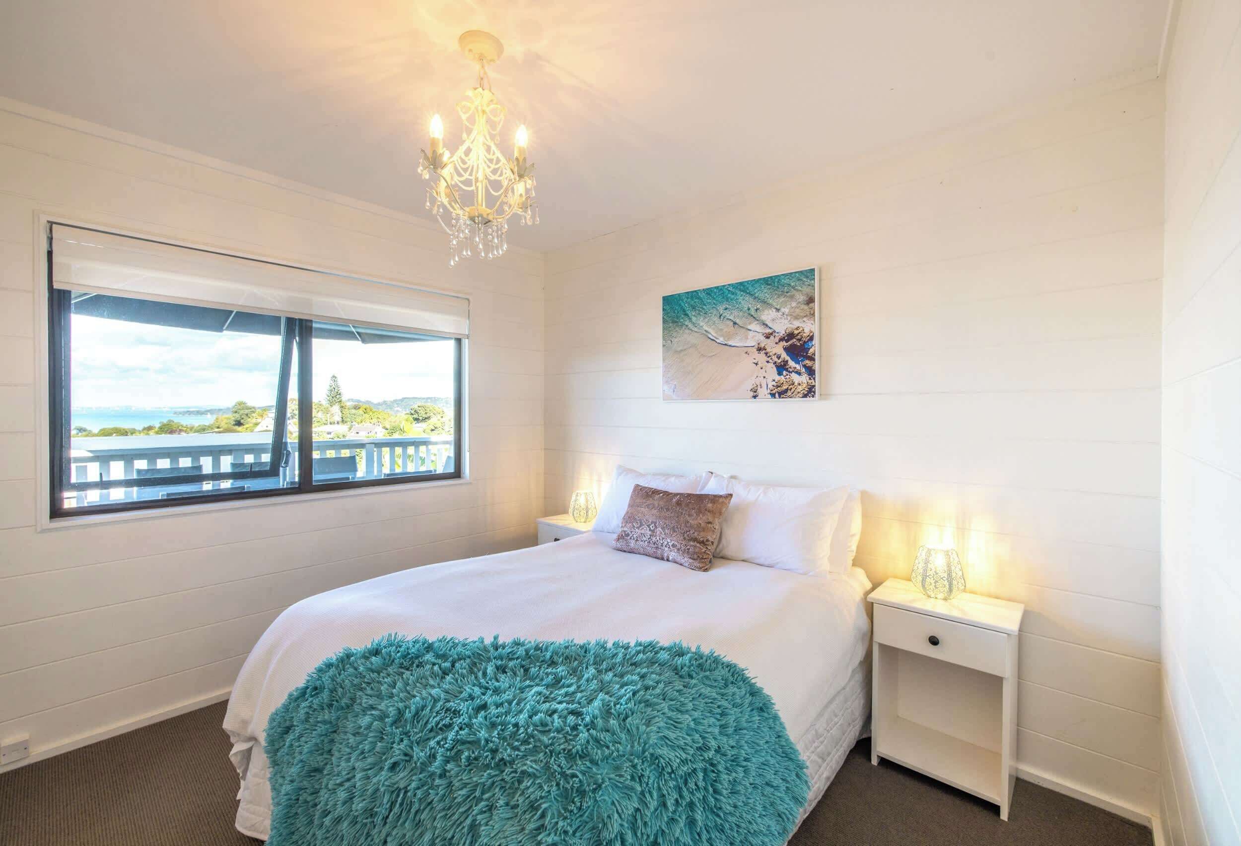 A bedroom with a bed, a window with a view, a chandelier, and a painting on the wall.